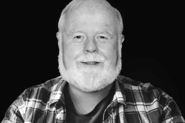 Portrait of an older man with a white beard smiling, wearing a plaid shirt against a dark background.
